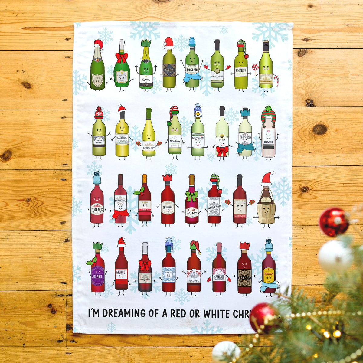 A white tea towel with a humorous Christmas illustration of wine bottles, laid flat on a wooden floor with holiday ornaments in the foreground.