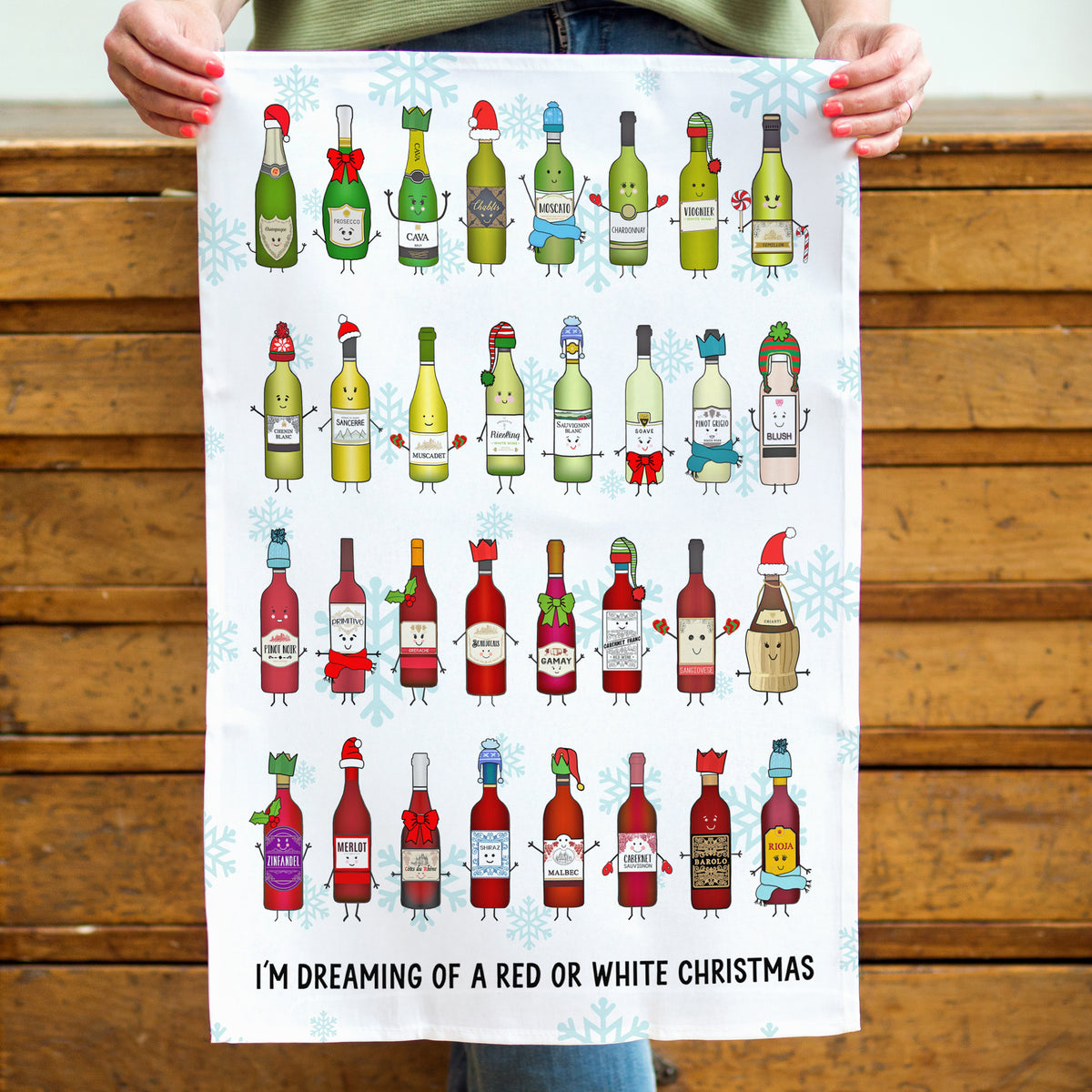 A person holds up a white rectangular print featuring illustrated wine bottles with Christmas-themed clothing and expressions, plus the text 'I'M DREAMING OF A RED OR WHITE CHRISTMAS' at the bottom. The background shows a wooden bench.