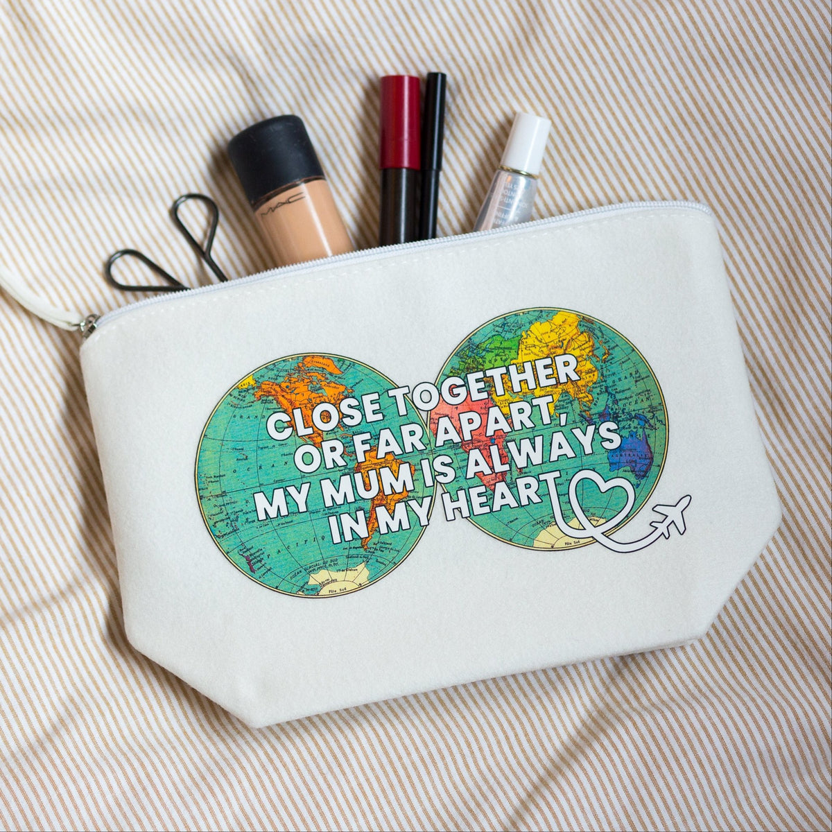 A white canvas cosmetic pouch with a colorful infinity symbol made of two globe-like circles and the text 'CLOSE TOGETHER OR FAR APART, MY MUM IS ALWAYS IN MY HEART'. Makeup items are tucked into the top zipper.