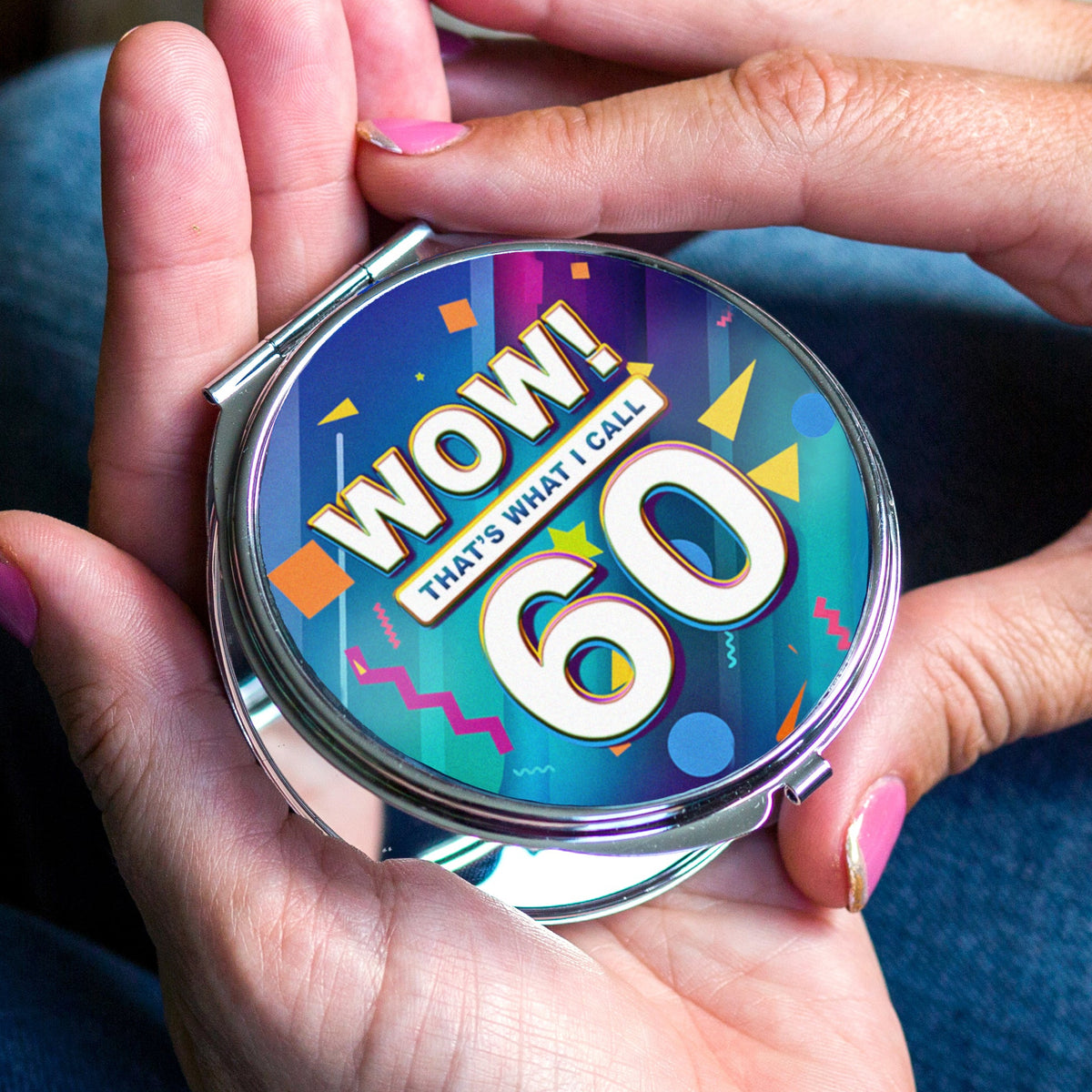 A person's hands holding a round silver compact mirror with a colorful, confetti-patterned screen that reads 'WOW! THAT'S WHAT I CALL 60'.