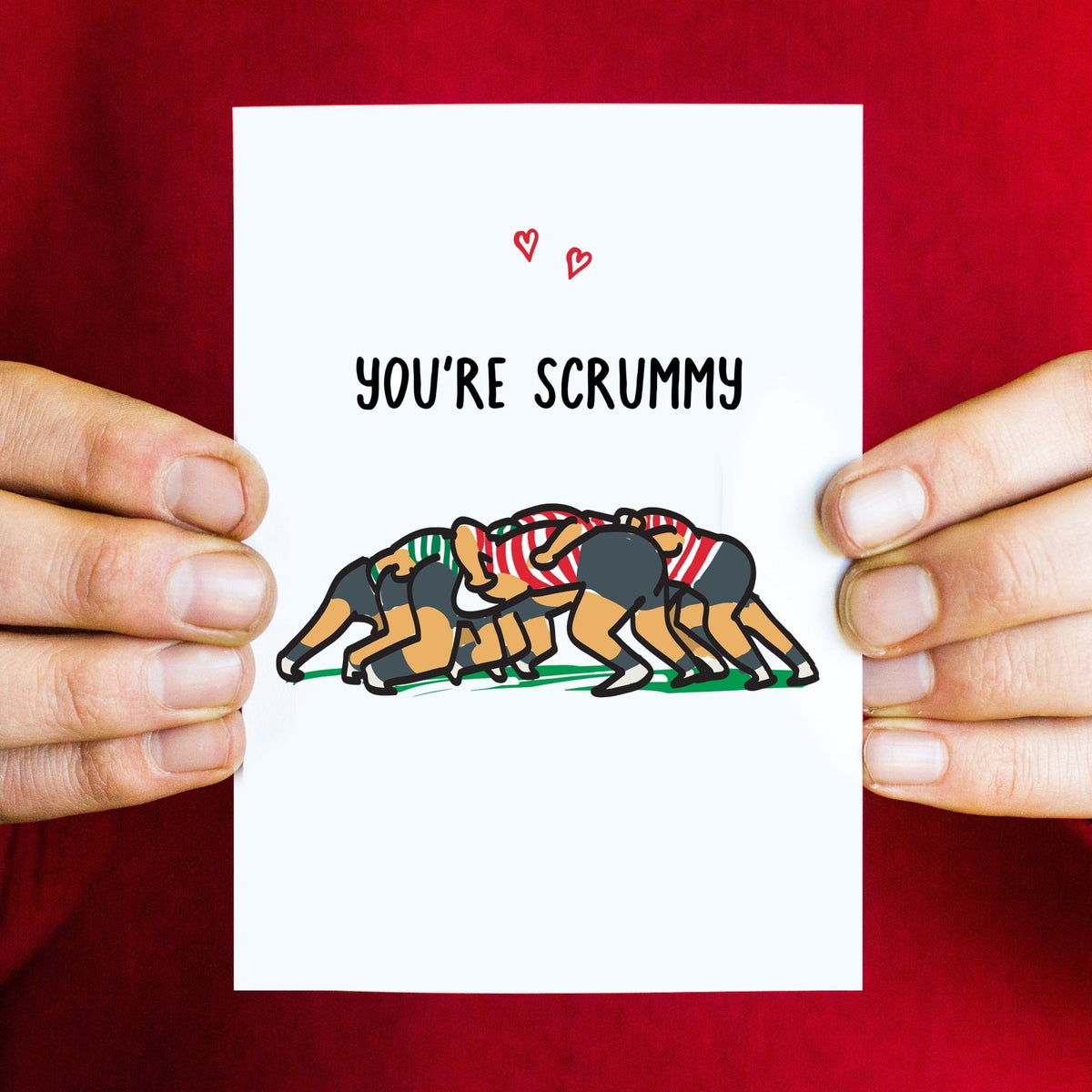a greeting card held in mans hands against a red jumper showing an illustration of a rugby scrum and the words 'You're Scrummy'