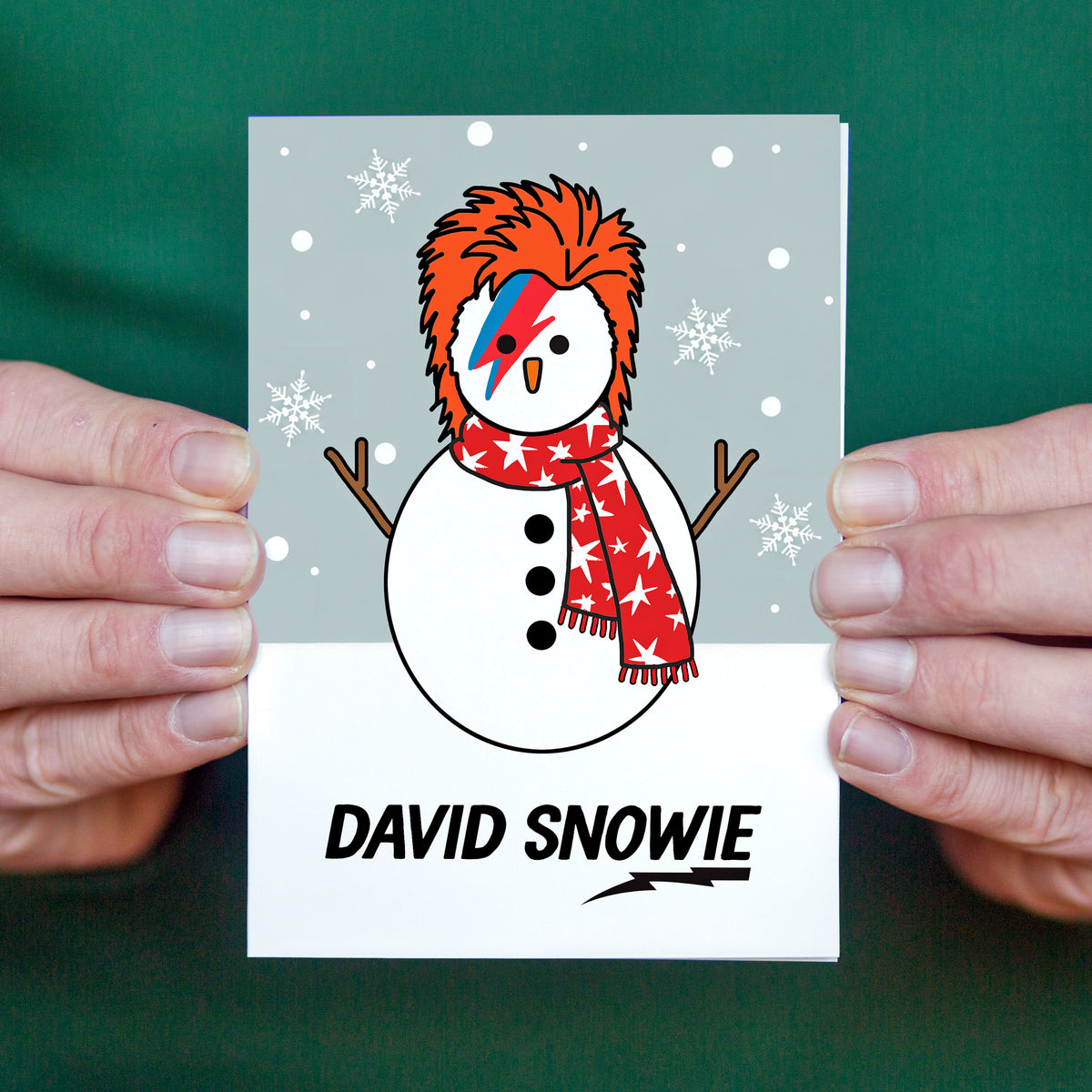 A custom greeting card featuring a snowman illustration with orange hair styled like David Bowie's lightning-embellished hair, a red star-patterned scarf, and 'DAVID SNOWIE' printed at the bottom. The card is held by two hands against a dark green background.