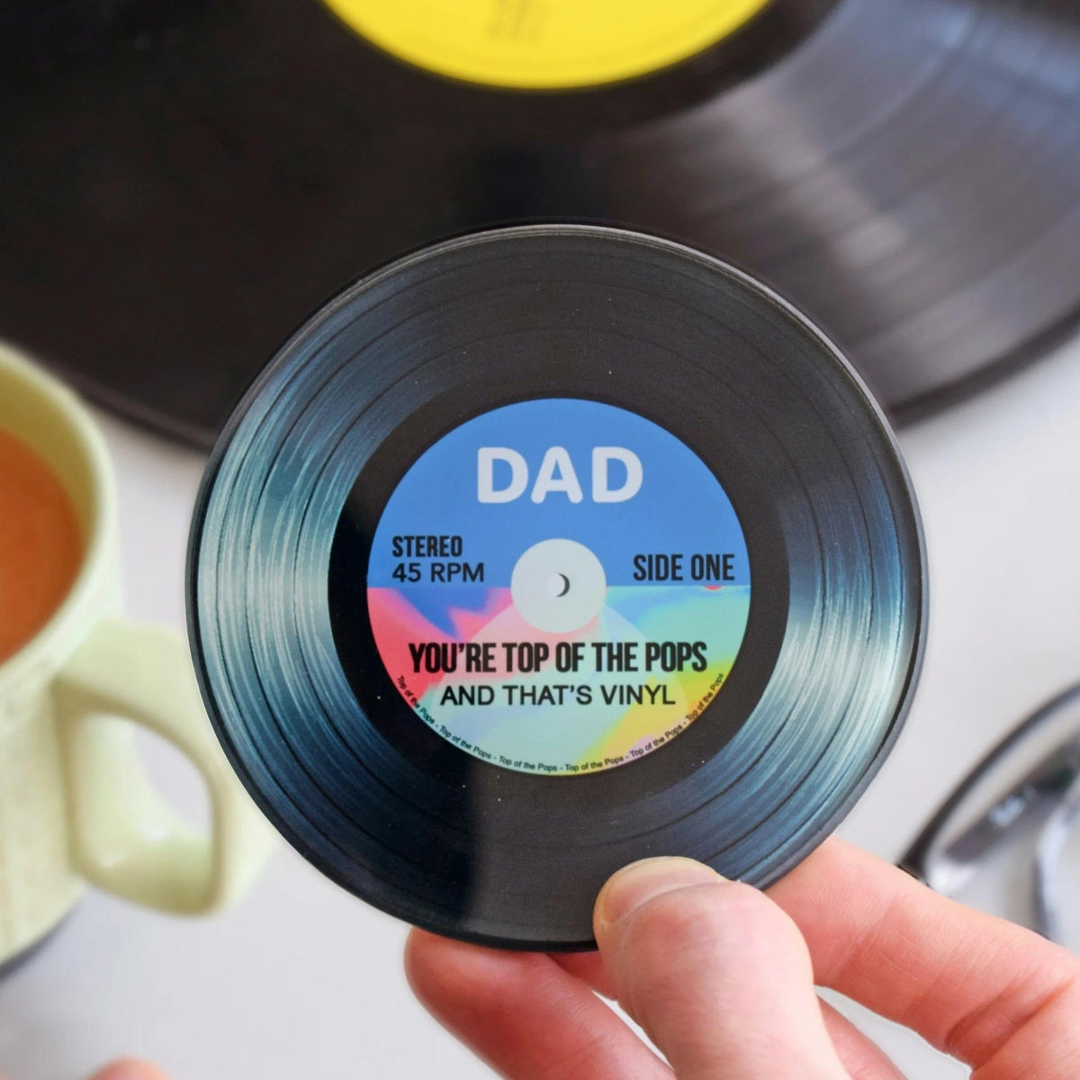 Small vinyl record with a 'DAD' label held by a hand, with a larger vinyl record in the background.