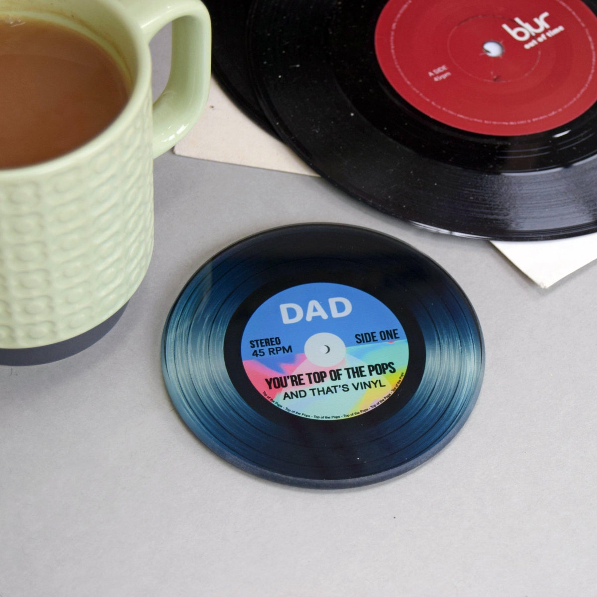 Vinyl record coaster with 'DAD' text next to a cup of coffee and records on a table.