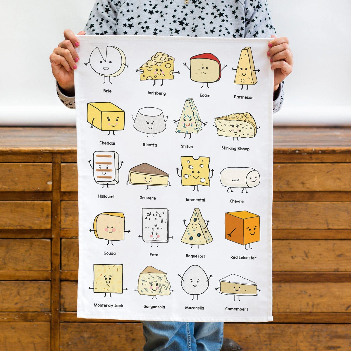 a white cotton tea towel featuring illustrations of all varieties of cheese held by a woman against a wooden background