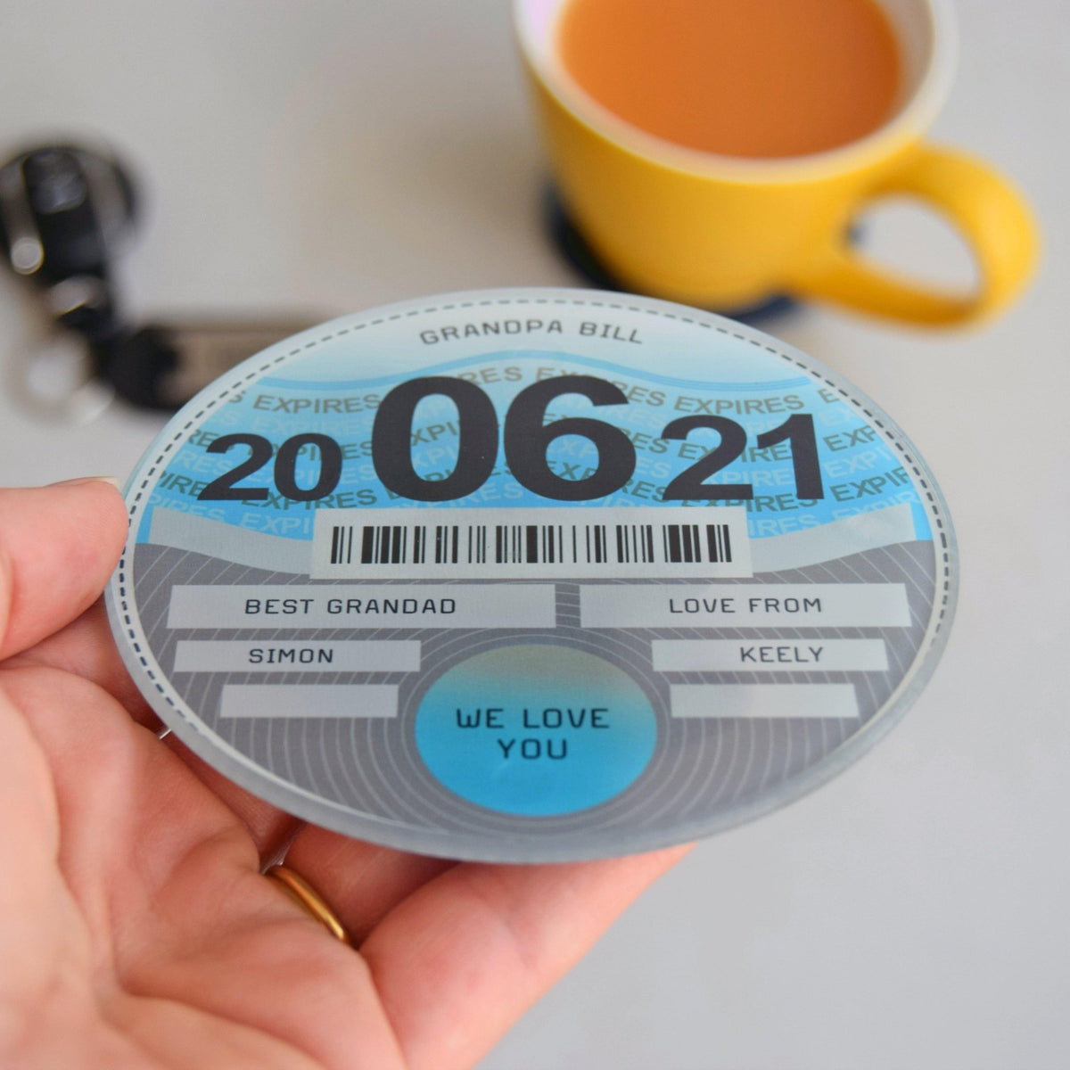 Round glass coaster with personalized message held by a hand, with a yellow mug in the background.