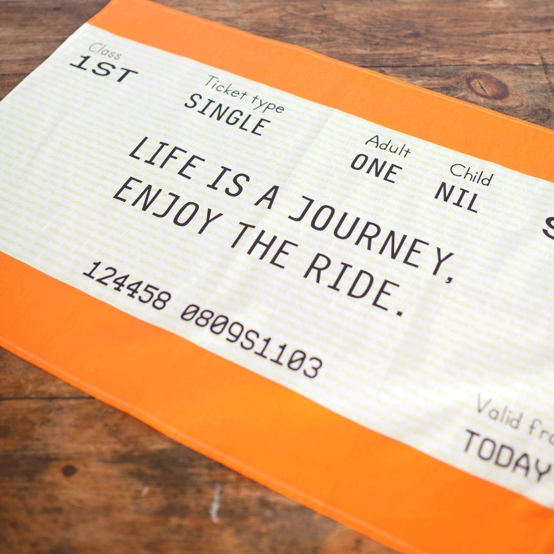 Train ticket tea towel with motivational message 'Life is a journey, enjoy the ride.' on a wooden surface